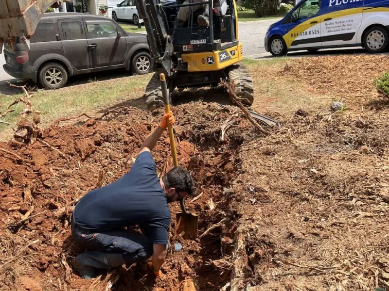Plumber digging out roots beside a mini excavator